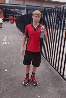 a young man in a red shirt and black shorts is holding a blue bottle of soda .