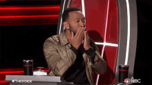 a man covering his mouth with his hands while sitting at a table that says the voice on it
