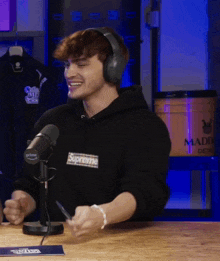 a man wearing headphones and a black supreme sweatshirt laughs in front of a microphone