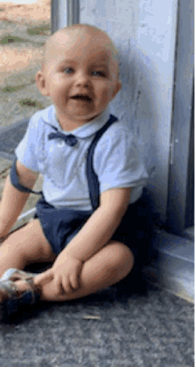 a baby wearing suspenders and a bow tie is sitting on the ground
