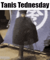 a man in a cape is walking in front of a blue flag with the words ' tanis tednesday ' on it .