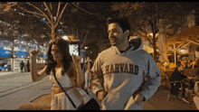 a man wearing a harvard sweatshirt walks down a street with a woman