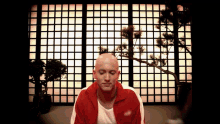 a bald man in a red jacket sits in front of a window with squares on it