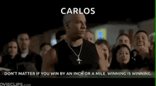 a man with a cross necklace is standing in front of a crowd of people and says `` carlos '' .