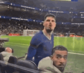 a man in a blue shirt is standing on a soccer field while another man looks on .