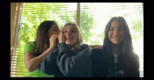 three young women are sitting next to each other in front of a window with blinds .