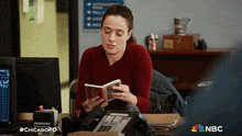 a woman sits at a desk reading a book with the nbc logo in the background