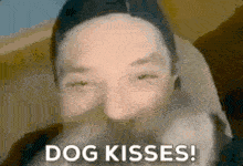 a man with a beard is holding a dog in front of his face and says `` dog kisses '' .