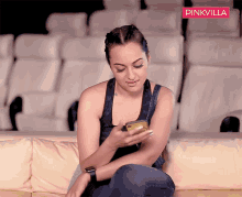 a woman sits on a couch looking at her phone with a pinkvilla logo in the corner