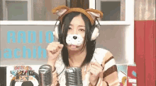a woman wearing headphones and a dog mask holds two microphones in front of a sign that says radio