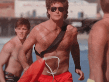 a shirtless man wearing red shorts and sunglasses walks on a beach