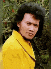 a man with curly hair and a beard is wearing a yellow shirt and looking at the camera .