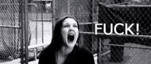 a black and white photo of a woman screaming in front of a chain link fence with the word fuck written on it .