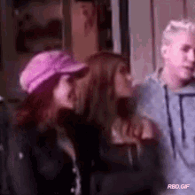 a man and two women are standing next to each other in a room . one of the women is wearing a pink hat .