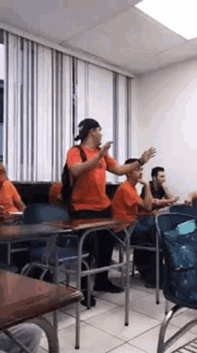 a man in an orange shirt is sitting at a desk in a classroom with other people .