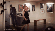 a woman in a black dress is dancing in a room with a man in a suit behind her