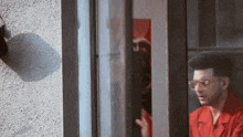 a man in a red shirt and glasses looks out of a window