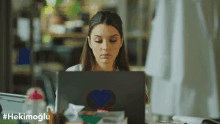 a woman sitting in front of a laptop with the hashtag #hekimoglu on the bottom right