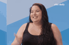 a woman in a black tank top is smiling in front of a blue wall with elite daily written on it