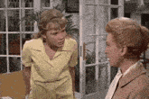 two women are standing next to each other in front of a window talking to each other .