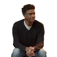 a man wearing a black sweater and jeans sits with his hands folded