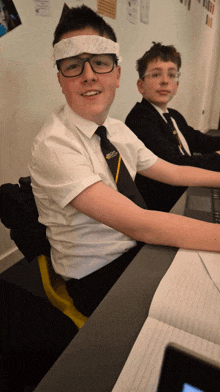 a boy wearing glasses and a bandana on his head is sitting at a desk next to another boy