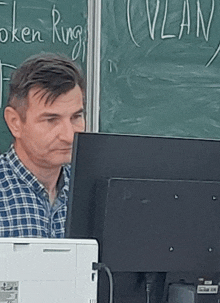 a man sitting in front of a computer with a token ring written on the blackboard behind him