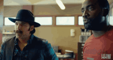 a man wearing a reebok shirt stands next to a man wearing a cowboy hat