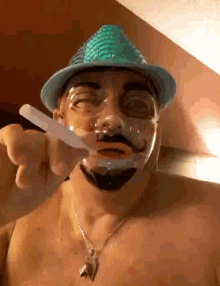 a shirtless man wearing a hat and a necklace is shaving his face with a razor ..