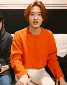 a young man wearing an orange sweater and white pants is sitting on a couch
