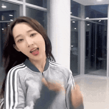 a woman in a gray jacket with white stripes on the sleeves is standing in a room with her tongue out .