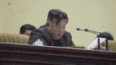 a man in a suit sits at a table with a microphone and a piece of paper in front of him .