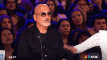 a bald man wearing sunglasses is speaking into a microphone in front of a crowd of people with a nbc logo in the background