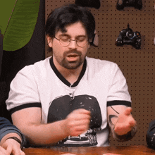 a man with glasses and a beard is sitting at a table with his hands out .