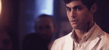 a man with a tattoo on his arm is wearing a white jacket and looking at the camera .