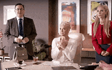 a man in a suit and tie is standing next to a woman in a white suit sitting at a table .