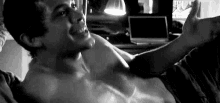 a shirtless man is laying on a bed with his arms outstretched and a laptop in the background .