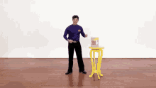 a man in a purple shirt is standing next to a yellow table with a box on it .