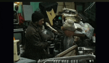 two men are standing in a room with a pile of junk including a microwave