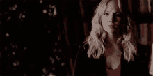 a woman with blonde hair and a red shirt is standing in a dark room looking at the camera .