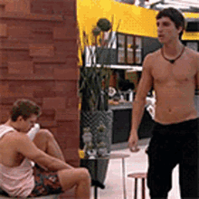 a shirtless man standing next to another shirtless man sitting on a bench