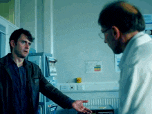 a man in a white lab coat talks to another man in a room