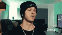 a man wearing a beanie and a necklace with the letter a on it looks at the camera
