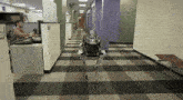a man sits at a desk in an office cubicle while a robot walks down a hallway
