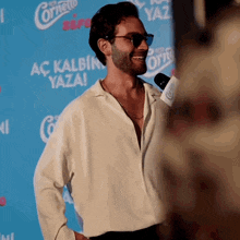 a man wearing sunglasses and a white shirt stands in front of a sign that says ac kalbin yaza