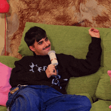 a man is sitting on a green couch holding a microphone and wearing a black sweater with the word ted on it