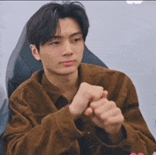 a young man in a brown jacket is sitting in a chair and making a heart with his hands .
