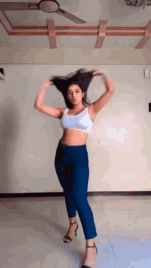 a woman in a white crop top and blue jeans is dancing in a room with a ceiling fan .