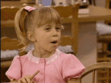 a little girl in a pink dress is sitting at a table holding a pencil and making a funny face .
