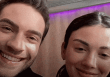 a man and a woman smile for the camera with purple lights behind them